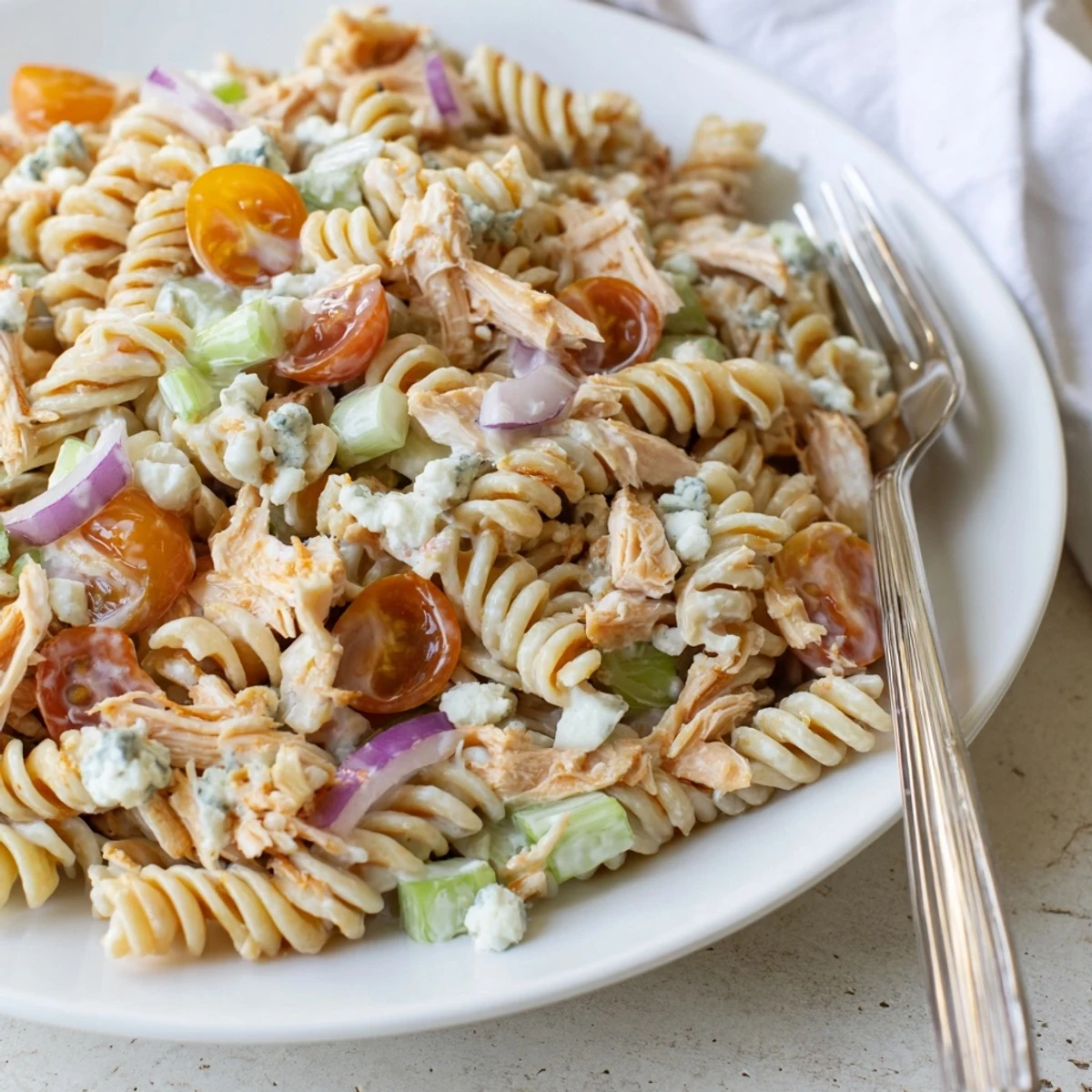 A vibrant bowl of Buffalo Chicken Pasta Salad featuring rotini pasta tossed with shredded buffalo chicken, diced celery, red bell peppers, and creamy ranch dressing, garnished with crumbled blue cheese and fresh chives.  