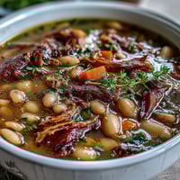 A steaming bowl of hearty ham and navy bean soup, rich with tender beans and smoky ham, garnished with fresh thyme.  